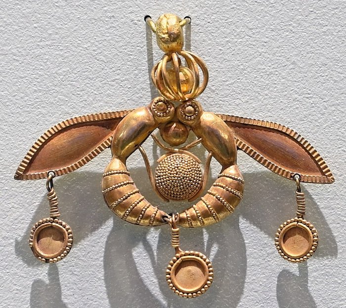 Archaeological Museum of Heraklion: Bee pendant excavated from the minoan Chrysolakkos necropolis at Malia, 1800-1700 BC, Isle of Crete, Greece. The pendant consists of two bees depositing a drop of honey in their honeycomb. They are holding the round honeycomb between their legs and the drop of honey in their mouths. On their heads the bees carry a delicate cage containing a golden bead. Small discs hang from their wings and stings. Size (width) 4.6 cm.